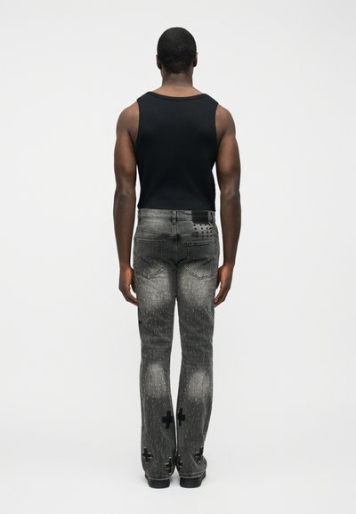 Άνδρας που φοράει μαύρο αμάνικο τοπ και γκρι ξεθωριασμένο τζιν με μοτίβα σταυρού στα πόδια, στέκεται με την πλάτη προς το μέρος μας σε απλό φόντο.