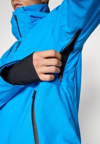 Zila mīkstā apģērba jaka ar augstu apkakli, ar rāvējslēdzēja kabatām un melniem akcentiem. Raksturīga gluda tekstūra un ergonomisks dizains.