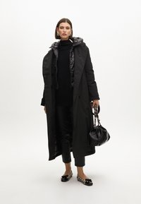 Černý dlouhý kabát s vycpanou šedou kapucí, vrstvený přes černý svetr a kalhoty. Černá kabelka s řetízkovým popruhem, tištěné ploché boty.