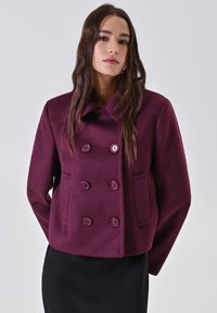 Cappotto doppiopetto in lana color bordeaux con ampio colletto, due tasche frontali e bottoni sporgenti. La texture è liscia, con una vestibilità sartoriale.