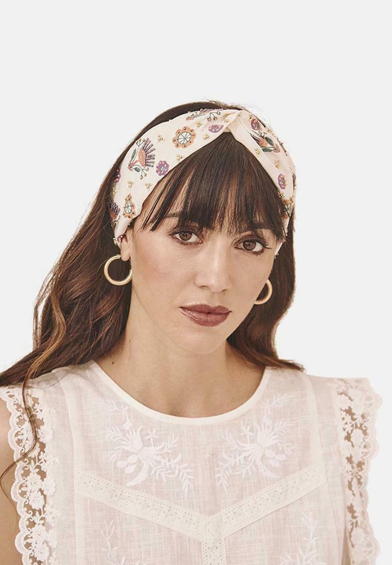 Diadema crema con patrón floral y diseño anudado. Textura suave. La modelo lleva una blusa blanca con adornos de encaje y cuello redondo.