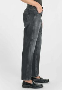 Jean en denim noir à coupe droite, avec taille mi-haute, poches latérales et décoloration subtile. Porté avec des mocassins noirs.