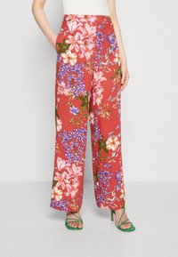 Bloemen wijde broek van rode stof, met roze en paarse bloemenpatronen, zijzakken en een relaxte pasvorm. Aangevuld met groene hakken.