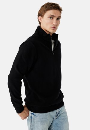 Young man wearing a black quarter-zip pullover and light blue jeans, standing with one hand in pocket against plain background.