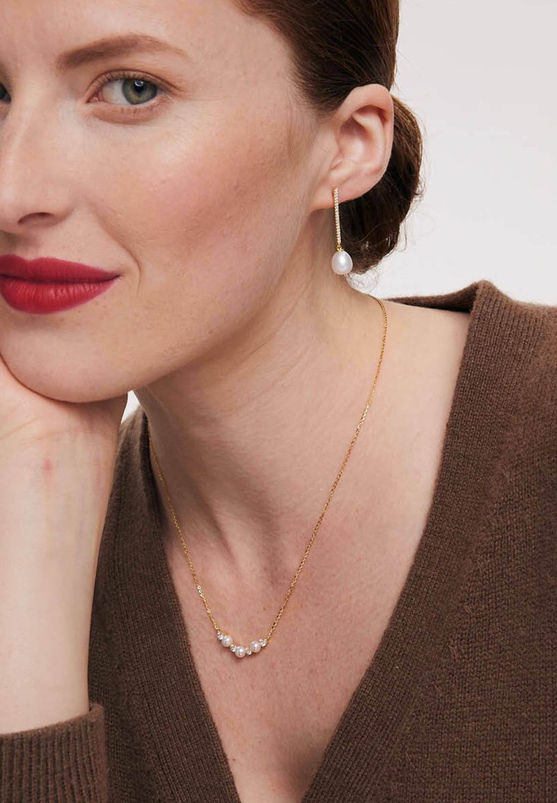 Złoty naszyjnik z łańcuszka z wieloma małymi perłami, noszony z złotą kolczykiem wiszącym z większą perłą. Widoczny dekolt brązowego swetra.