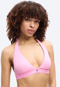 Γυναίκα με σγουρά μαλλιά που φοράει ροζ μπικίνι halter με το λογότυπο Tommy Hilfiger, σε απλό λευκό φόντο.