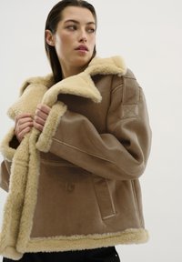 Giacca in shearling marrone con colletto beige peloso, fronte con bottoni e maniche oversize. La texture è morbida, con una finitura esterna liscia.
