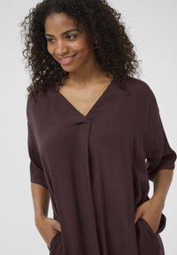 Top corto bordeaux con scollatura a V e tasche laterali. Tessuto morbido e liscio, vestibilità comoda con silhouette drappeggiata.