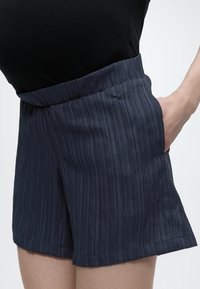 Μπλε ναυτικό ριμπ υφασμάτινο σορτς με ελαστική μέση, φορεμένο με μαύρο τοπ, χέρι να ακουμπά στην πλαϊνή τσέπη.