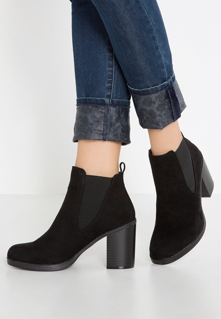 Bottines en daim noir avec panneaux latéraux élastiques, un bout arrondi et un talon épais. Portées avec un jean denim foncé retroussé orné d'une bordure à motif.