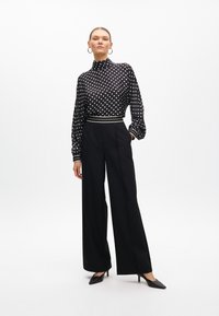 Black polka dot blouse with high neck and long sleeves, paired with high-waisted black wide-leg trousers. Black pointed heels complete the outfit.