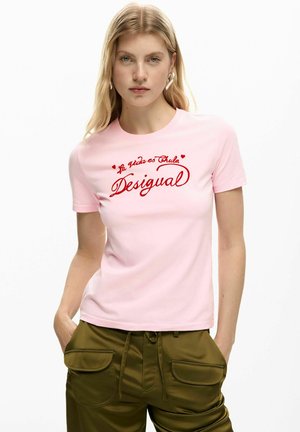 Donna bionda che indossa una maglietta rosa chiaro con scritta rossa "La vida es Chula Desigual" e pantaloni verde oliva con grandi tasche frontali.