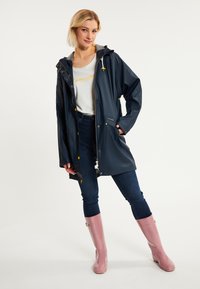 Marinblå vattentät regnjacka med huva, guldfärgade tryckknappar och en framficka, bärs över en ljusgrå topp och mörka jeans, med rosa gummistövlar.