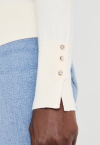Pull crème avec des poignets côtelés comportant trois boutons décoratifs. Le pull a une fente au bas des poignets, associé à un jean bleu.