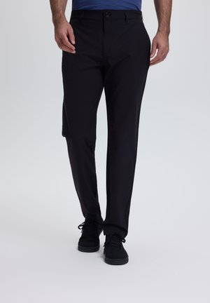 Homme portant un pantalon noir à jambes droites et des chaussures noires à lacets, debout devant un fond clair uni.