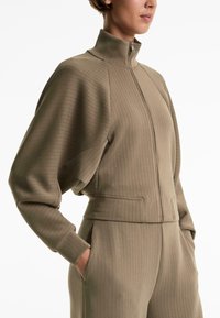 Giacca zip-up marrone chiaro con texture a coste, colletto alto e maniche ampie. Presenta tasche laterali e una vita aderente.