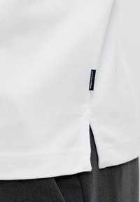 Gros plan sur l'ourlet d'une chemise blanche avec fente latérale et petite étiquette noire indiquant "JACK & JONES" au-dessus d'un pantalon gris foncé.