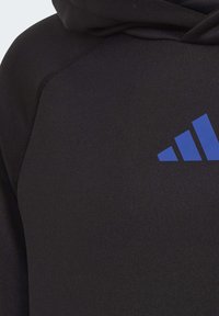 Svart hoodie med en texturerad tyg, med en framträdande blå Adidas-logotyp i nedre högra hörnet och dragsko vid huvan.