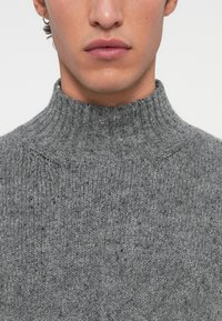 Nahaufnahme des unteren Gesichts und des Halses einer Person, die einen grauen gestrickten Rollkragenpullover und einen kleinen silbernen Creolenohrring trägt.