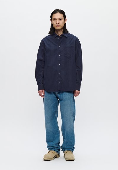 Camisa azul marino de botones con rayas verticales, bolsillo en el pecho, combinada con jeans de denim azul claro y calzado beige.