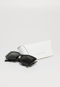 Ochelari de soare negri cu rame unghiulare stau lângă o carcasă rectangulară albă cu text reliefat. Lentilele sunt întunecate și de formă ovală.