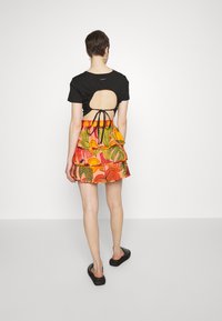 Černý top s krátkým rukávem a výřezem na zádech, zakončený uzlem, v kombinaci s barevnou mini sukní s vrstveným designem a botanickými vzory. Černá obuv.