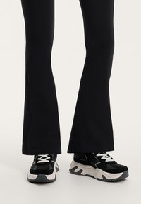 Fekete harangszárú leggings fekete és fehér sportcipővel párosítva. A sportcipő fekete felsőrésszel rendelkezik, texturált részletekkel, és fehér talppal, szürke kiegészítőkkel.