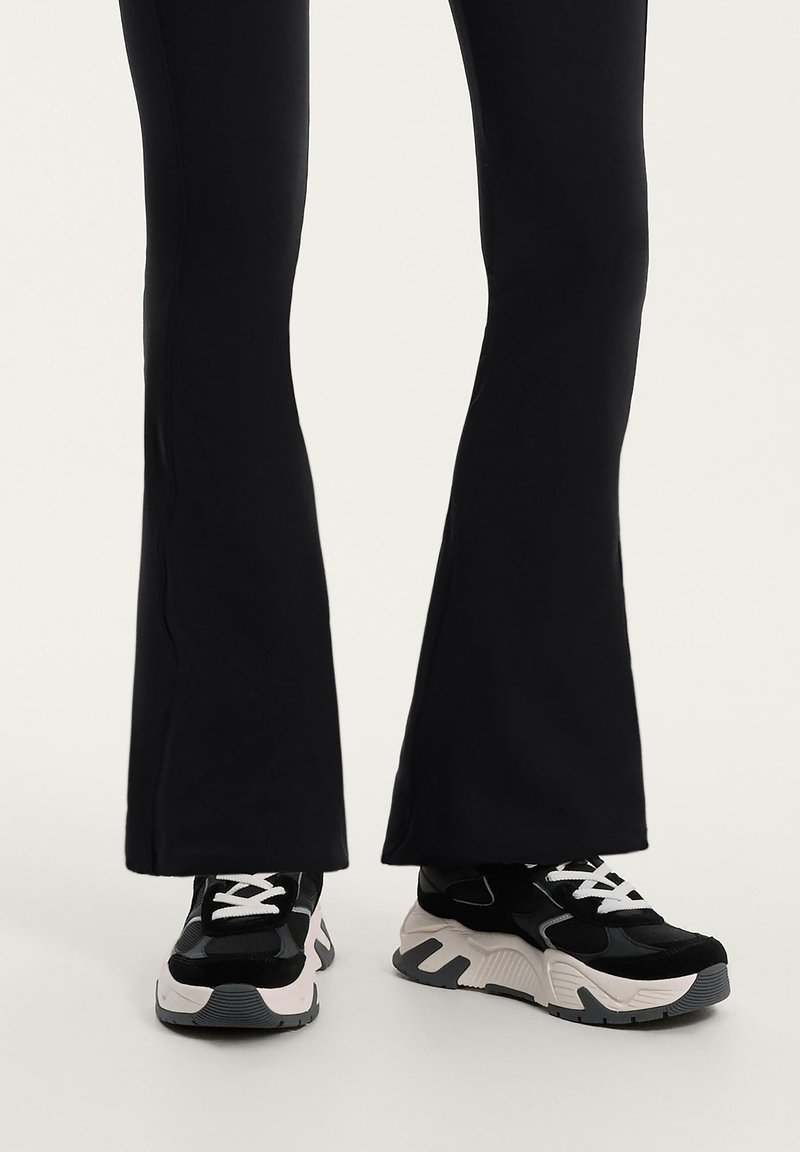 Fekete harangszárú leggings fekete és fehér sportcipővel párosítva. A sportcipő fekete felsőrésszel rendelkezik, texturált részletekkel, és fehér talppal, szürke kiegészítőkkel.