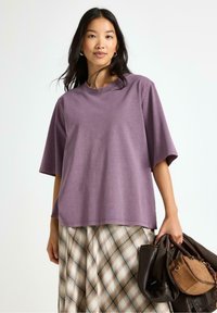 Lilla oversized t-shirt i blødt stof med rund halsudskæring, parret med en ternet nederdel. Modellen holder en brun håndtaske.