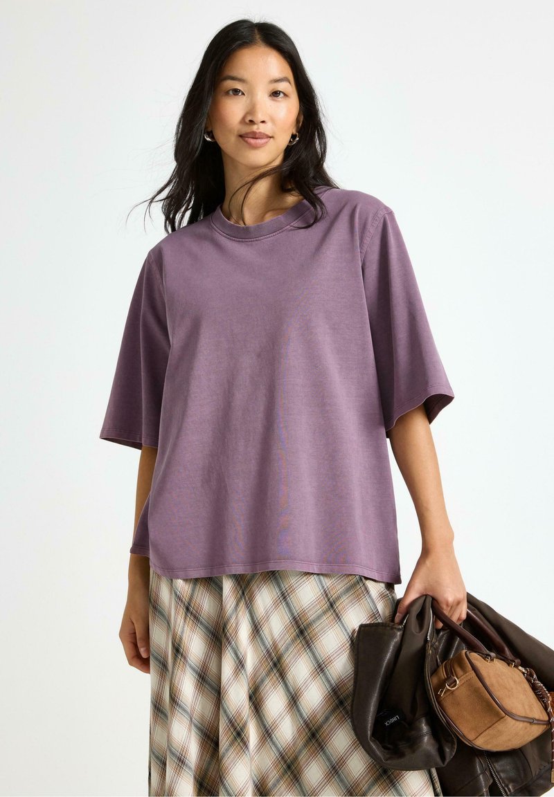 Lilla oversized t-shirt i blødt stof med rund halsudskæring, parret med en ternet nederdel. Modellen holder en brun håndtaske.