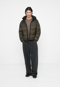 Chaqueta acolchada en verde oliva oscuro con cierre de cremallera frontal, puños acanalados y capucha desmontable, combinada con pantalones de pierna ancha negros.