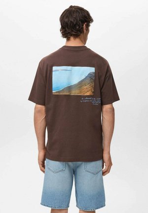 T-shirt in cotone marrone con una grafica quadrata di un paesaggio costiero sul retro, caratterizzata da acqua blu e tonalità terrose. Vestibilità casual.
