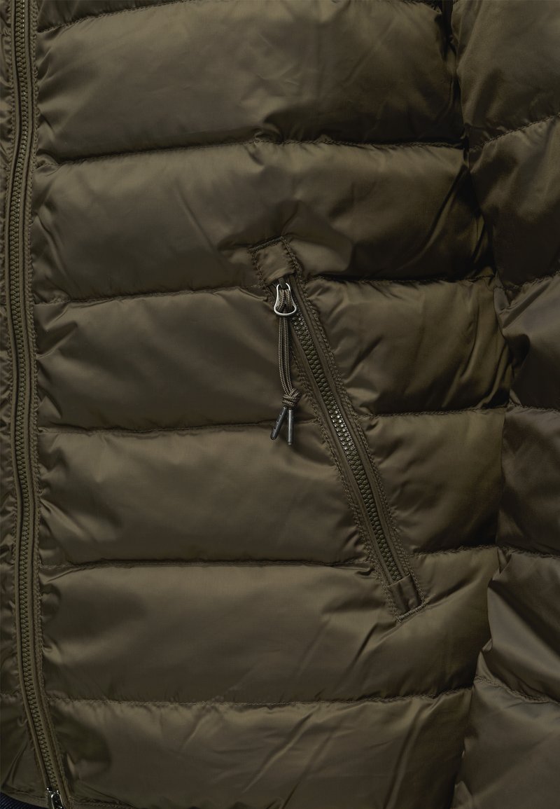 Nahaufnahme einer olivgrünen Steppjacke mit horizontalen Nähten und einer Reißverschlusstasche mit Textilreißverschluss-Zuglasche.
