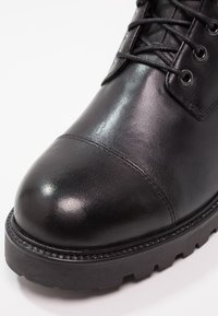 Svart läderstövel med en glänsande yta, rundad tå, dekorativ sömnad och en tjock, mönstrad gummisul. Snörningsdesign med metallhål.