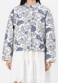 Kurteczka pikowana z białym tłem i niebieskim motywem paisley, z okrągłym kołnierzem, zapięciem na guziki i długimi rękawami.