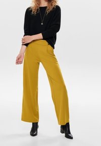 Žluté široké kalhoty s hladkým povrchem, spárované s černým svetrem s dlouhým rukávem. Černé kotníkové boty dotvářejí outfit.