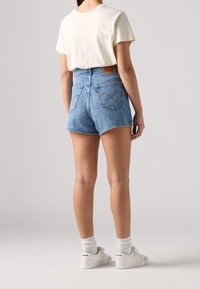 Shorts de mezclilla azul de tiro alto con cinco bolsillos, dobladillo deshilachado y parche de cuero en la parte posterior. Combinados con una camiseta blanca y zapatillas blancas.