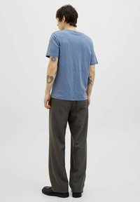 Kék pamut gallér nélküli póló rövid ujjal, laza illeszkedésű sötétszürke nadrággal és fekete cipővel párosítva. A karokon látható tetoválásokkal rendelkezik.