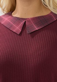Närbild av en ribbad bordeauxfärgad topp med en rosa rutig spetskrage på en person med blont hår.
