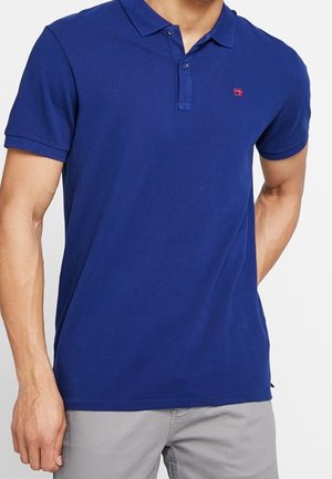 Mężczyzna ubrany w gładką niebieską koszulkę polo z krótkim rękawem, z małym czerwonym logo na piersi, oraz jasnoszare spodnie.