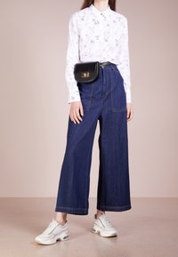 Camicia bianca con fantasie, abbinata a jeans in denim blu a vita alta e a gamba larga. Borsa in vita nera e sneakers bianche con dettagli a contrasto.