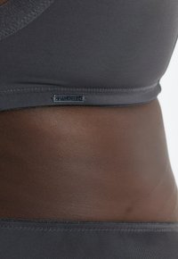 Szary biustonosz wykonany z gładkiej tkaniny; posiada płaską taśmę w pasie z małą metką marki. Tekstura wydaje się subtelna i gładka na tle koloru skóry.