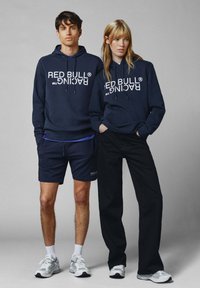 Sudaderas de la Marina con el estampado "RED BULL RACING", combinadas con pantalones de pierna ancha negros y pantalones cortos azul marino. Los modelos llevan zapatillas blancas con acentos grises.