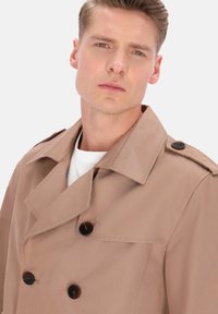 Lyse beige trenchcoat med dobbeltradet design, store knapper og brede revers, lavet af et tekstureret stof.