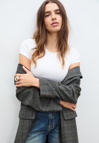 Grauwe visgraatjas over een witte T-shirt, met blauwe jeans. Opvallende gouden ring aan de hand, casual pose, gestructureerde stof.