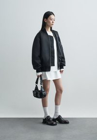 Μαύρο μπουφάν, άσπρο φόρεμα, μαύρη τσάντα με καρφιά, άσπρες κάλτσες και λαμπερά μαύρα παπούτσια με πόρπη, σε ουδέτερο φόντο.