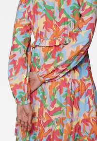 Robe à volants colorée en tissu léger avec des motifs abstraits en orange, rose, vert et bleu. Comprend une taille froncée et des manches longues.