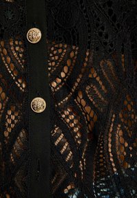 Blusa nera in pizzo con motivi floreali intricati, una pattina dei bottoni nera e un dettaglio in bottone dorato. Texture semi-trasparente evidente.