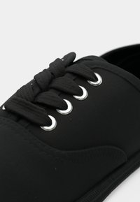 Gros plan d'une basket noire avec de grosses lacets noirs passant à travers quatre œillets argentés sur une tige en tissu lisse.