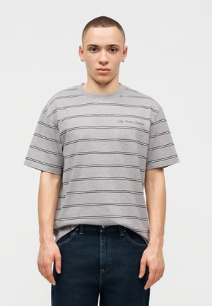 Noor mees lühikeste juustega, seljas halli triibulise tikiga t-särk ja tumedad teksad, seismas lihtsa heleda tausta ees.